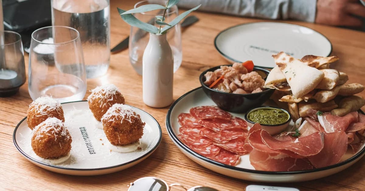 Malaga tapas platter: croquetas, jamón serrano, charcuterie, flatbread, pesto, prawns on stylish plates with water glasses and greenery.