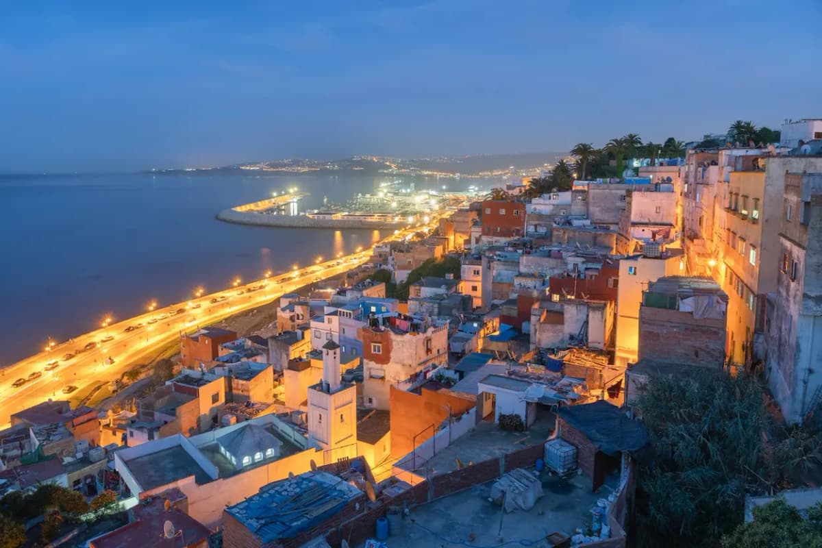 Aerial view of Tangier medina rooftops and port at dusk, with the Bay of Tangier and illuminated coastal promenade below
