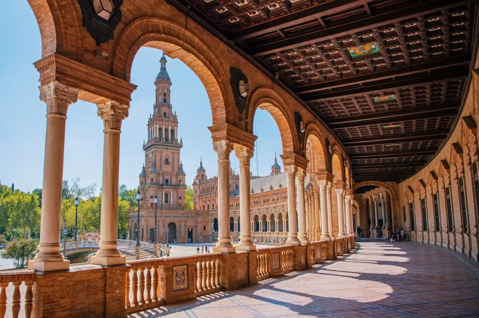 Plaza de España in Seville, Spain — the vast semicircular building with provincial tile alcoves reflected in the canal, tiled bridges and gondola boats under a blue Andalusian sky