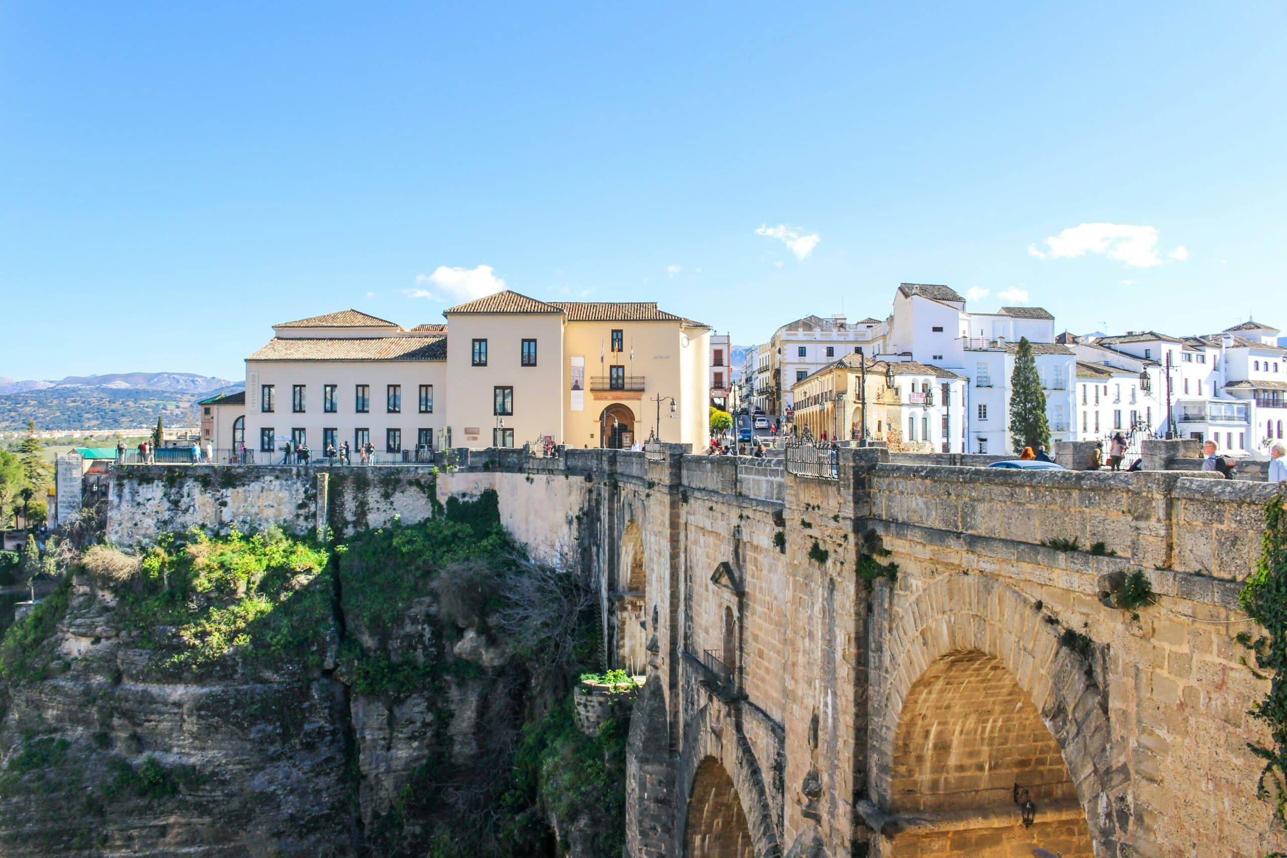 View of Ronda, Spain: the stone arch bridge Puente Nuevo spanning the deep El Tajo gorge, with white and pastel buildings lining the cliff under a bright blue sky.
