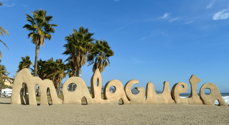Sculpted “Malagueta” sign on the sandy beachfront of La Malagueta in Malaga, Spain, with palm trees and a clear blue sky in the background.