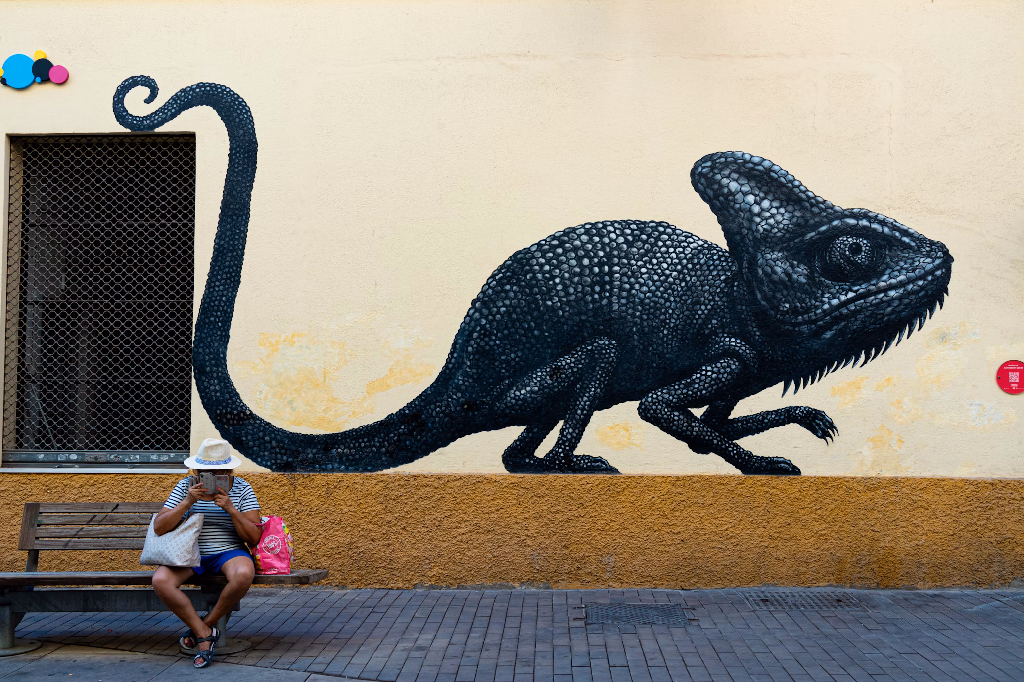Street art mural of a black chameleon on a wall in Malaga’s Soho district, with a man sitting on a bench below, reading.